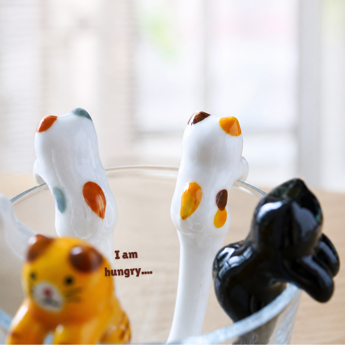A close-up of ceramic cat-shaped spoons in a clear glass bowl, including white, black, yellow, and orange cats, with a soft-focus indoor background and the text 'I am hungry.