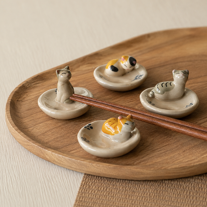 Four small ceramic cat-shaped chopstick rests arranged on a round wooden tray, with two resting under a pair of wooden chopsticks. The cat figures are painted in soft neutral tones with one cat in yellow and orange and small blue floral details.