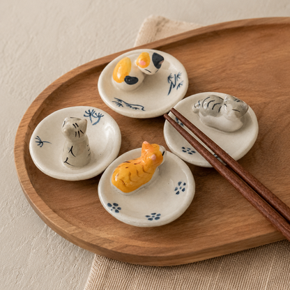 Four small white ceramic dish holders shaped like cat animals, arranged on a round wooden tray with a pair of chopsticks. The holders feature hand-painted blue floral details and colorful animal figures in gray, orange, yellow, and black.