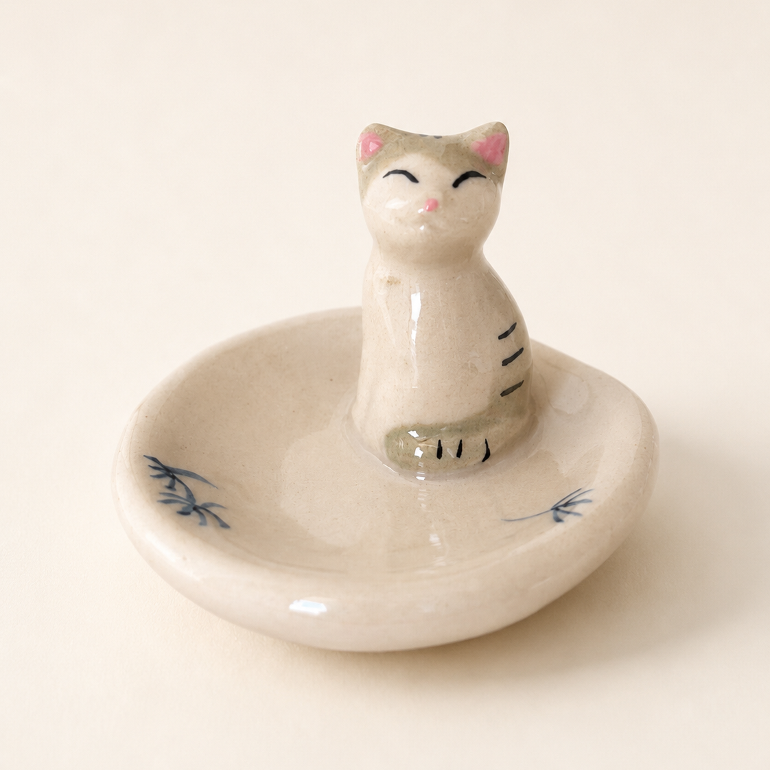 A ceramic cat-shaped chopstick rests, ring holder and tray set on a warm cream background. The glossy off-white ceramic cat figurine sits on a matching shallow dish decorated with small blue floral details.