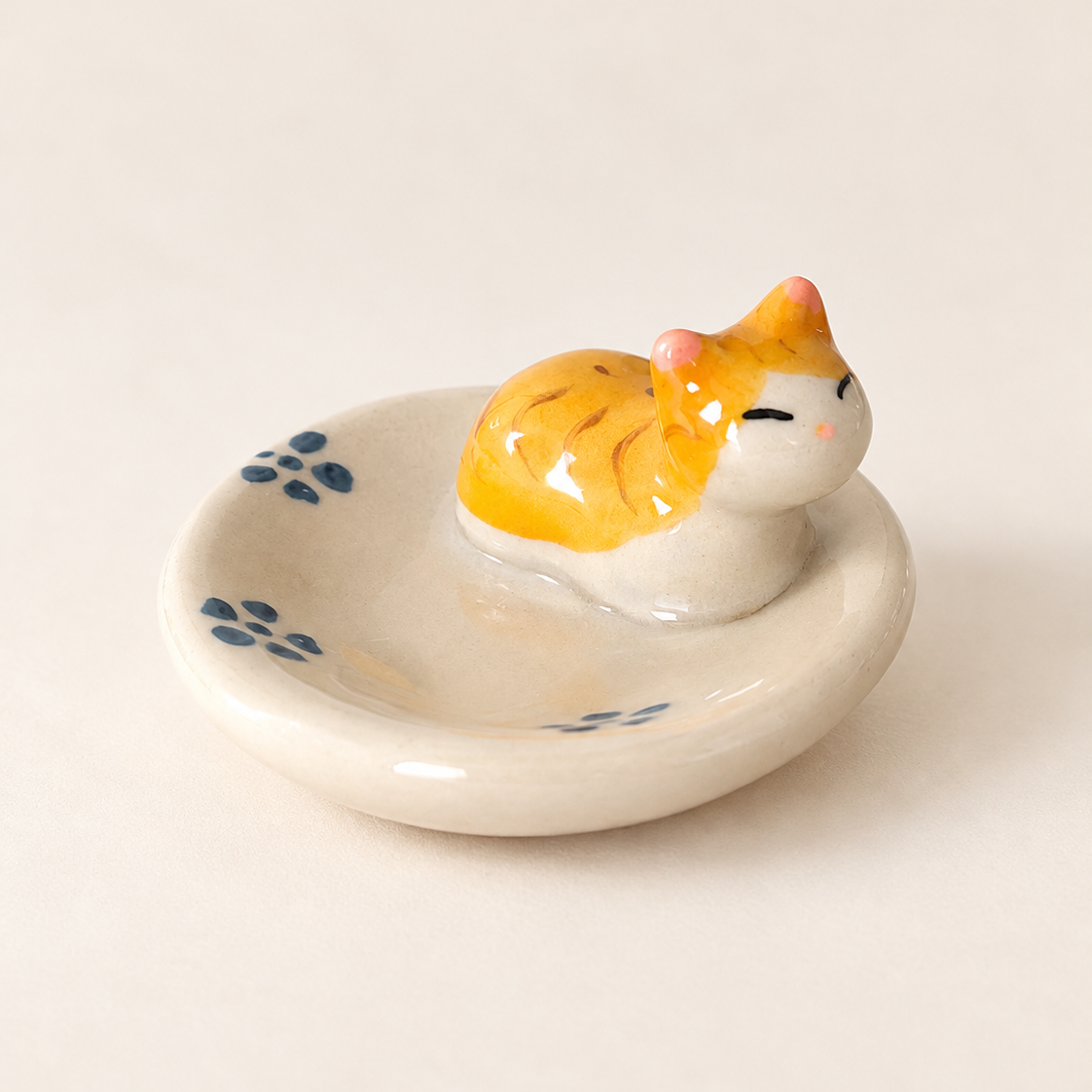 A ceramic cat-shaped utensil holder and chopstick rest with a small orange and white cat figurine sitting on a round off-white dish decorated with blue floral motifs, set against a plain light beige background.