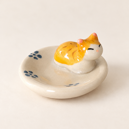 A ceramic cat-shaped utensil holder and chopstick rest with a small orange and white cat figurine sitting on a round off-white dish decorated with blue floral motifs, set against a plain light beige background.