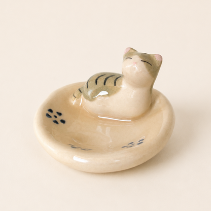 A small ceramic cat-shaped chopstick rest or utensil or brushes, ring holder with a cream-colored glaze, featuring a stylized cat sitting in a shallow bowl with subtle blue painted details and a green stripe pattern on its back.