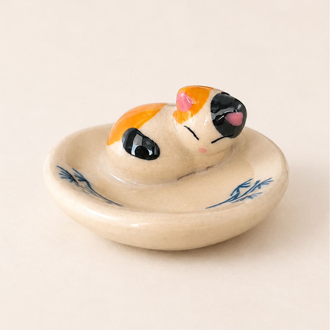 A ceramic cat-shaped chopstick rest or ring holder with a cream-colored glaze, blue leaf patterns on the rim, and a small orange, black, and pink cat figurine resting inside.