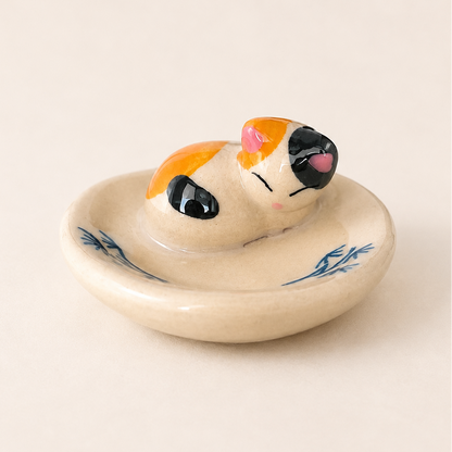A ceramic cat-shaped chopstick rest or ring holder with a cream-colored glaze, blue leaf patterns on the rim, and a small orange, black, and pink cat figurine resting inside.