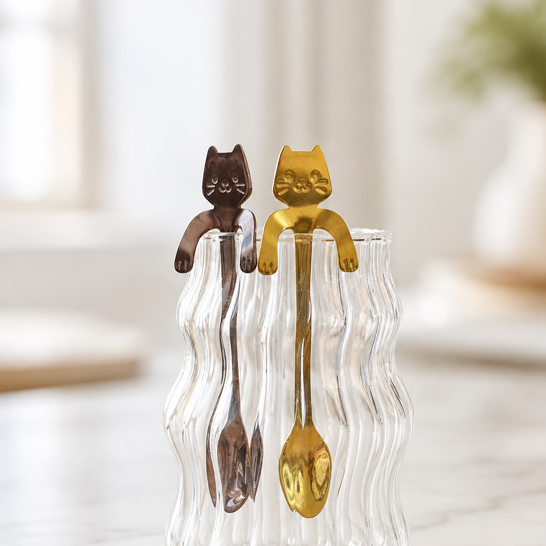 Two cat-shaped spoons, one gold and one dark brown, hanging from a wavy clear glass container on a light marble surface with a softly blurred indoor background.