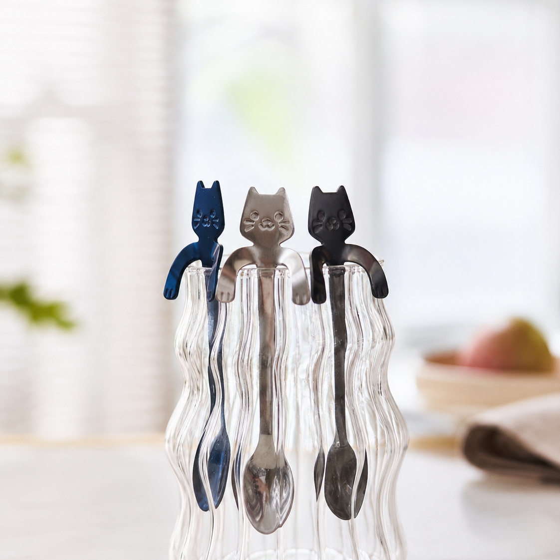 Three decorative spoons with cat-shaped metal spoon in blue, silver, and black, displayed in a wavy clear glass holder on a light marble countertop with a softly blurred kitchen background.