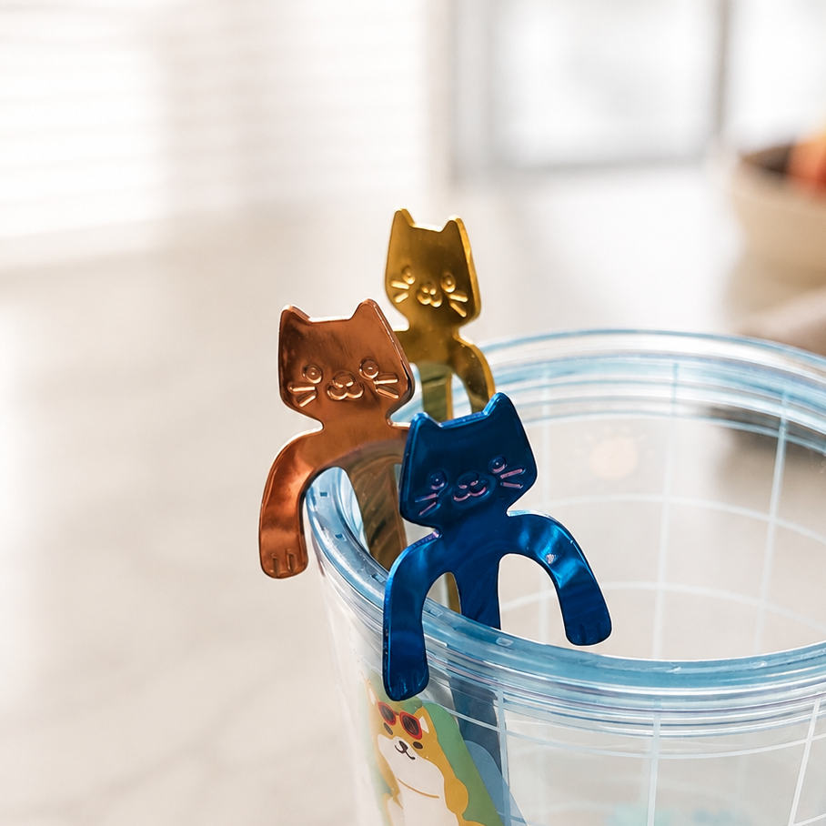 A clear plastic tumbler with a blue rim and dog-themed graphics, featuring three metallic cat-shaped metal spoon in gold, brown, and blue resting on the rim.