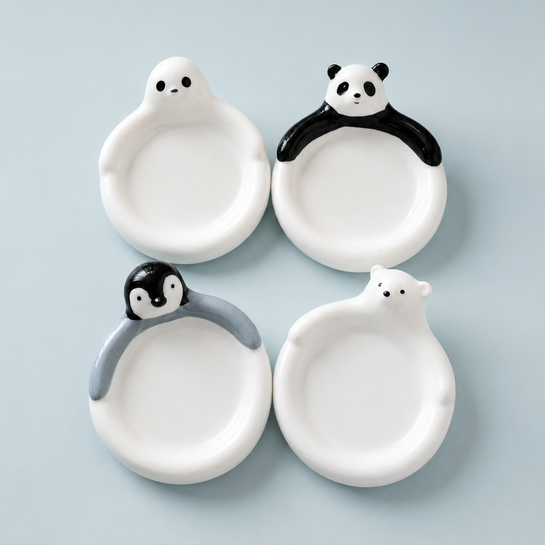 Four small white ceramic plates shaped like cartoon animals, arranged in a 2x2 grid on a light blue background. The top-left plate is seal-shaped, the top-right is panda-shaped, the bottom-left is penguin-shaped, and the bottom-right is polar bear-shaped.
