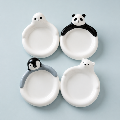 Four small white ceramic plates shaped like cartoon animals, arranged in a 2x2 grid on a light blue background. The top-left plate is seal-shaped, the top-right is panda-shaped, the bottom-left is penguin-shaped, and the bottom-right is polar bear-shaped.