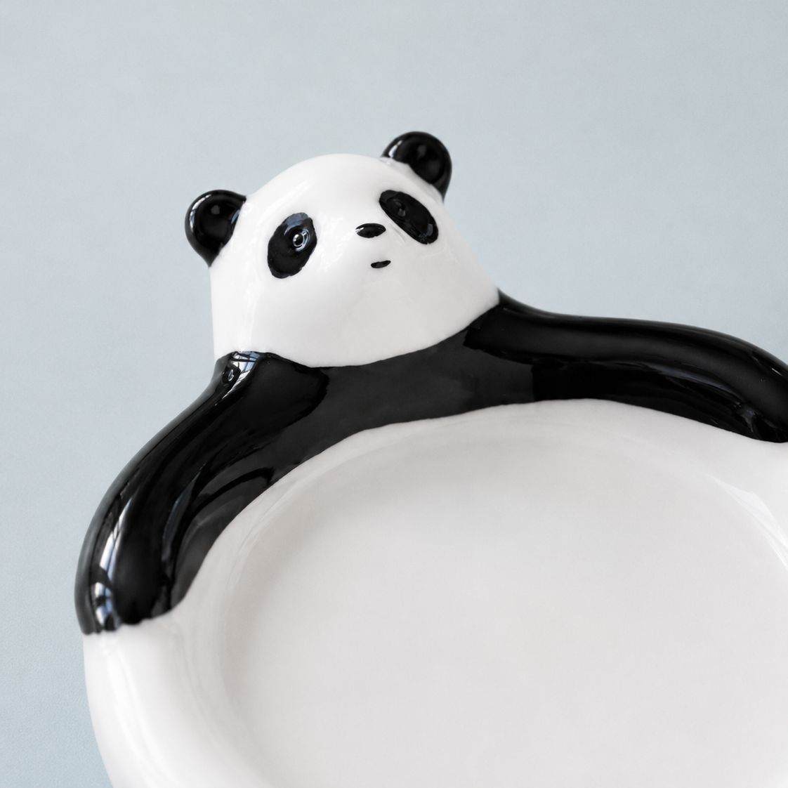 A close-up of a glossy black and white ceramic plate shaped like a panda bear sitting behind it, set against a solid light gray background.