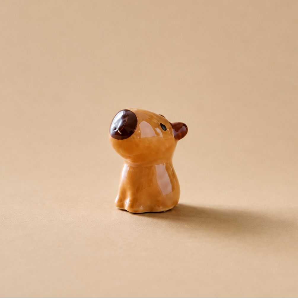 A small glazed ceramic animal  figurine, shaped like a cartoon capybara with a warm orange-brown body, dark brown nose and ears, and small green eyes, standing on a beige background.