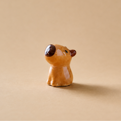 A small glazed ceramic animal  figurine, shaped like a cartoon capybara with a warm orange-brown body, dark brown nose and ears, and small green eyes, standing on a beige background.