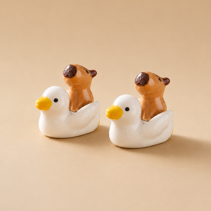 Two small ceramic duck-shaped figurines with brown capybara figures riding on their backs, set against a solid beige background.