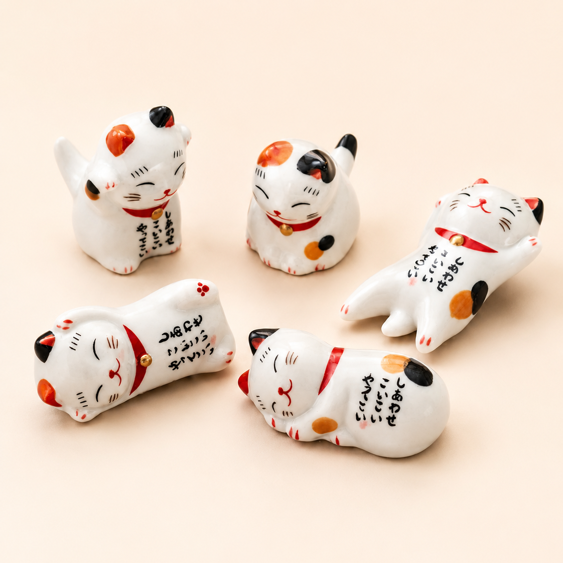 Five white ceramic Maneki-neko lucky cat figurines with black, orange, and red painted details, arranged on a solid pale peach background.