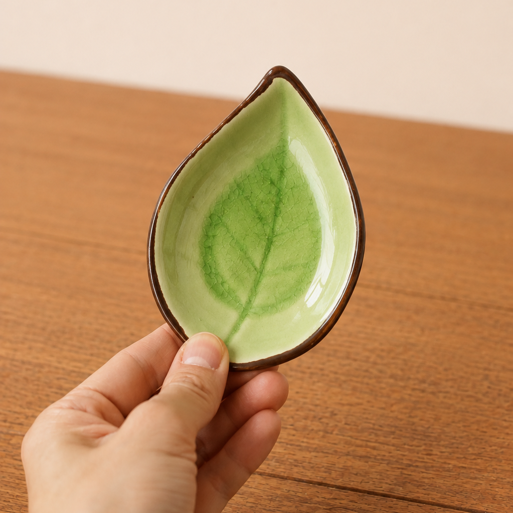 A hand holding a small leaf-shaped ceramic dish with a glossy light green interior featuring a darker green leaf pattern and a dark brown rim, set against a wooden table and beige wall background.