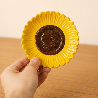 A hand holding a small yellow ceramic plate shaped like a sunflower, featuring a dark brown textured center, set against a warm beige wall and wooden tabletop.
