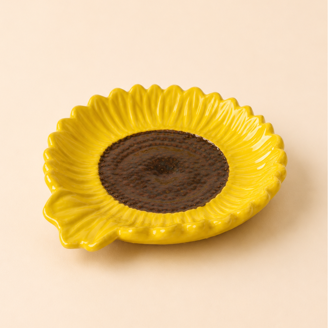 A yellow ceramic dish shaped like a sunflower, featuring textured yellow petals and a dark brown center, set against a solid beige background.