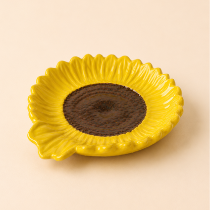 A yellow ceramic dish shaped like a sunflower, featuring textured yellow petals and a dark brown center, set against a solid beige background.