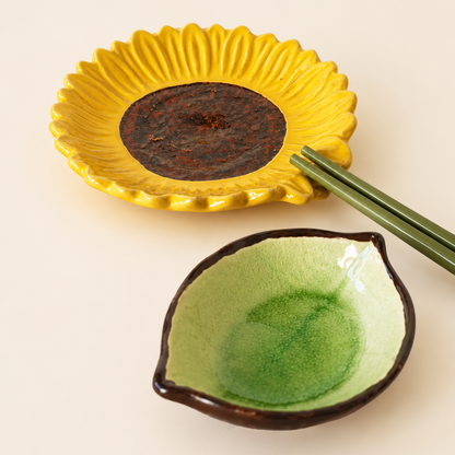 A ceramic dish set featuring a yellow sunflower-shaped plate holding a dark brown sauce, paired with green chopsticks, and a leaf-shaped green glazed bowl below, on a warm off-white background.
