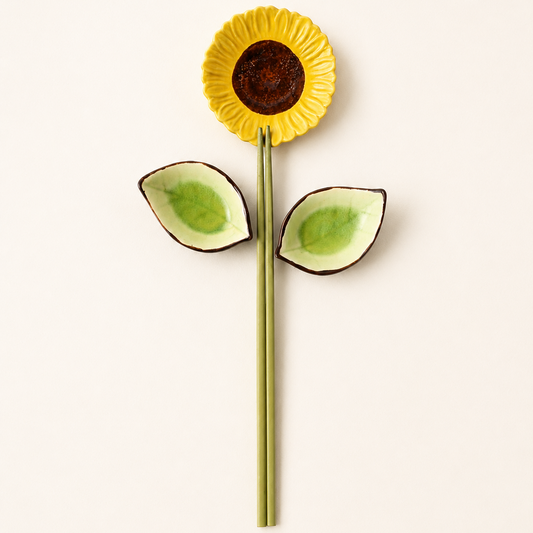 A decorative flower-shaped dish or spoon holder with a yellow sunflower head, dark brown center, and two green leaves on a stem, photographed on a plain light background.