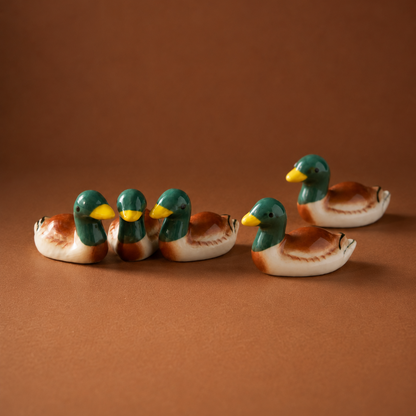 Four small ceramic mallard duck figurines and chopstick rests or utensil holder arranged on a smooth brown studio surface against a matching brown background.