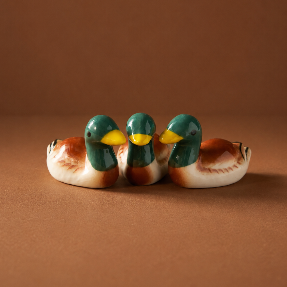 Three small ceramic mallard duck figurines and chopstick rests or utensil holder with glossy green heads, yellow beaks, and brown-and-white bodies, arranged side by side on a warm brown studio background.
