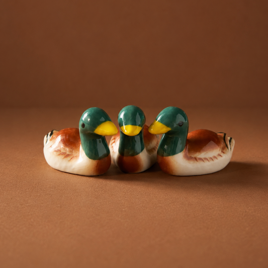 Three small ceramic mallard duck figurines and chopstick rests or utensil holder with glossy green heads, yellow beaks, and brown-and-white bodies, arranged side by side on a warm brown studio background.
