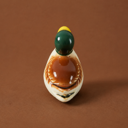 A small ceramic duck figurine and chopstick rests or utensil holder with a glossy brown body, cream-colored head and lower body, a dark green head, and a yellow beak, set against a solid brown background.
