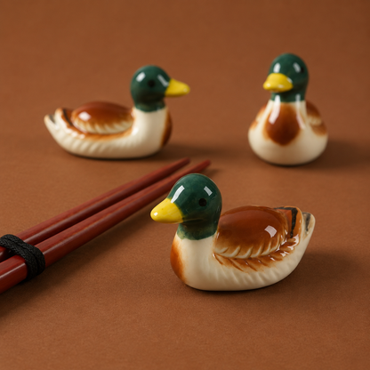 Three ceramic duck figurines and chopstick rests or utensil holder in mallard-style glaze with green heads, yellow beaks, and brown-and-cream bodies, arranged on a warm brown surface with a pair of red chopsticks in the foreground.
