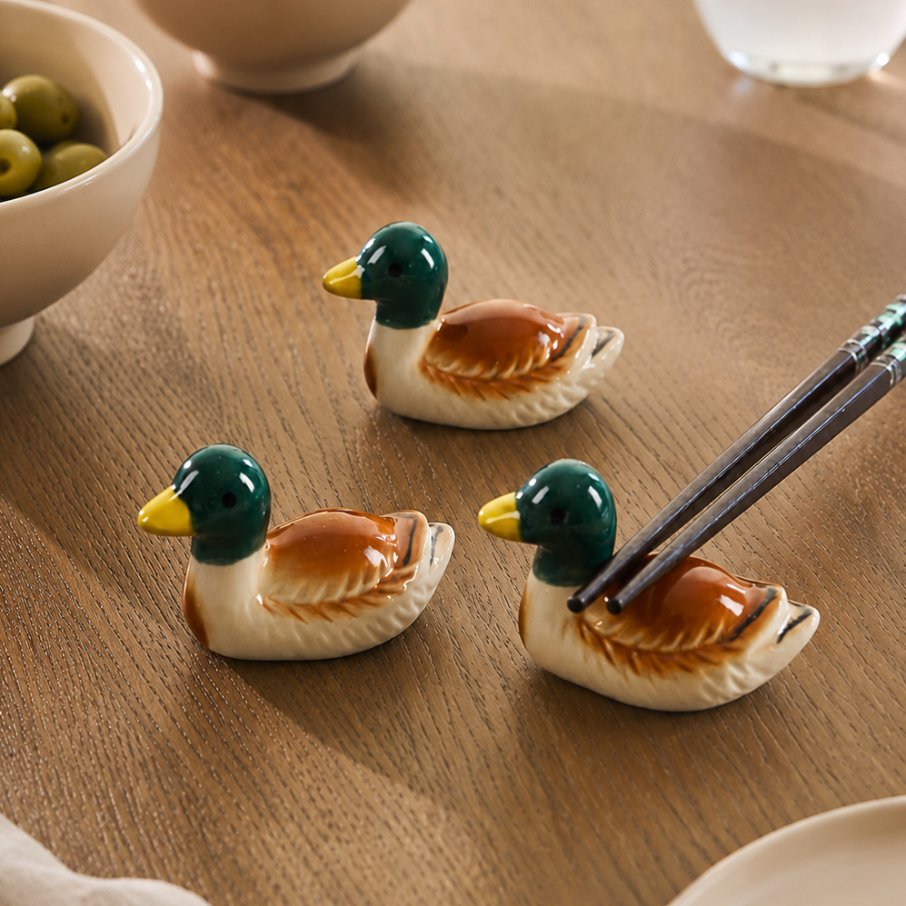 Three ceramic duck-shaped chopstick holders on a wooden table, with two holding dark chopsticks. The scene includes bowls of olives and nuts, a glass, and a plate in a styled dining setting.