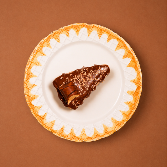 A single chocolate-covered pastry or dessert piece sits centered on a white plate with a waffle-like orange border, set against a solid warm brown background.