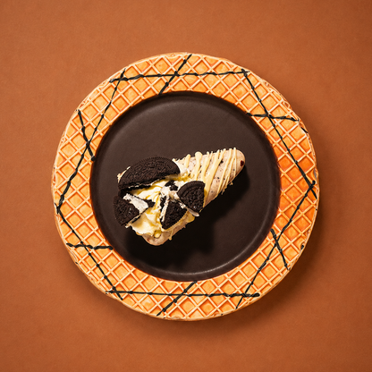 A dessert slice with cream, chocolate drizzle, and Oreo cookie pieces, presented on a dark plate with an orange waffle-patterned rim, set against a solid brown background.