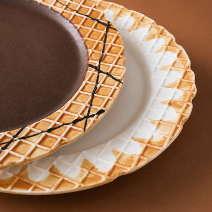 Three ceramic plates with a waffle-ice cream design, stacked diagonally on a warm brown background. The top plate is dark brown with black crisscross lines, the middle plate is white with a waffle pattern, and the bottom plate shows a white and brown waffle pattern with a scalloped edge.