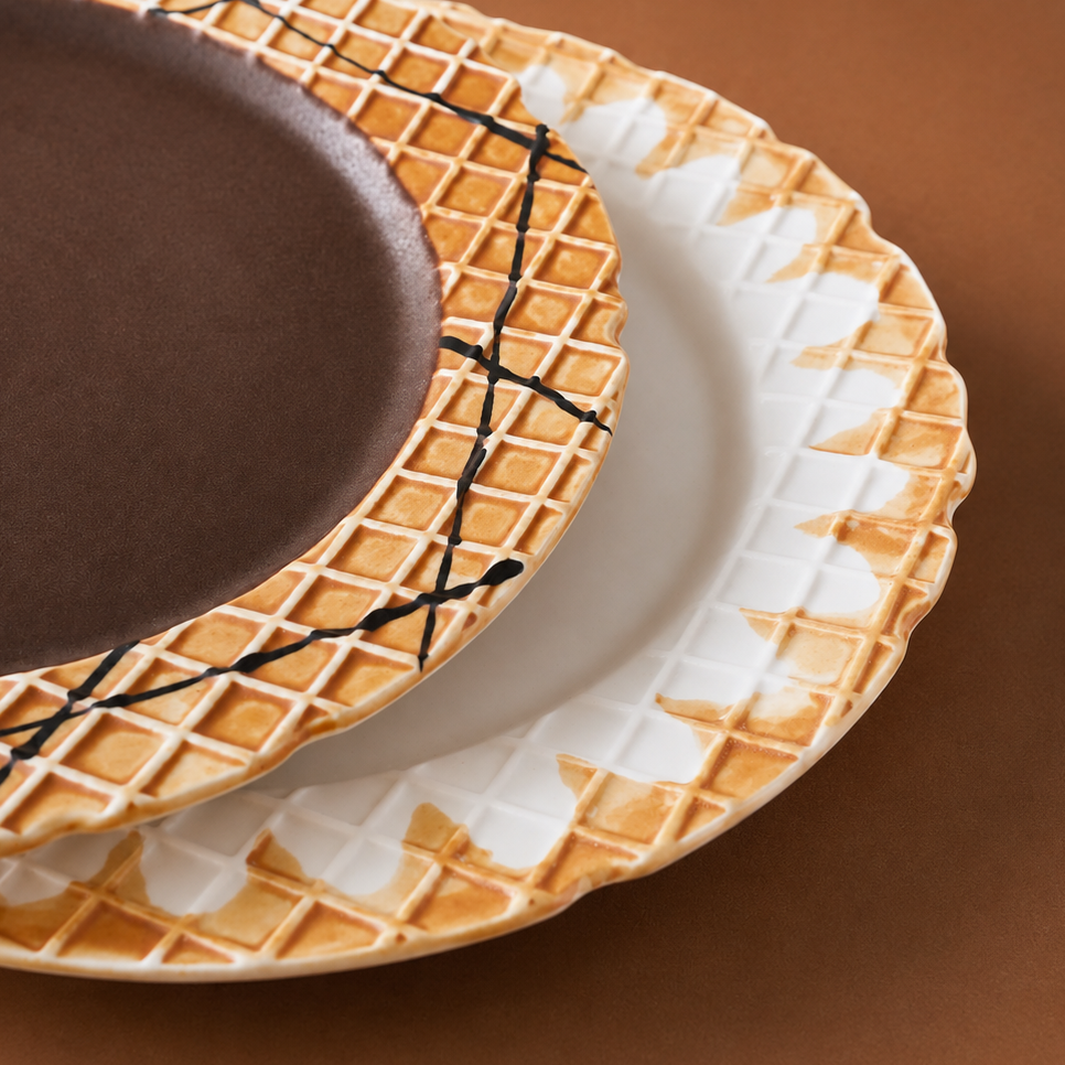 Three ceramic plates with a waffle-ice cream design, stacked diagonally on a warm brown background. The top plate is dark brown with black crisscross lines, the middle plate is white with a waffle pattern, and the bottom plate shows a white and brown waffle pattern with a scalloped edge.
