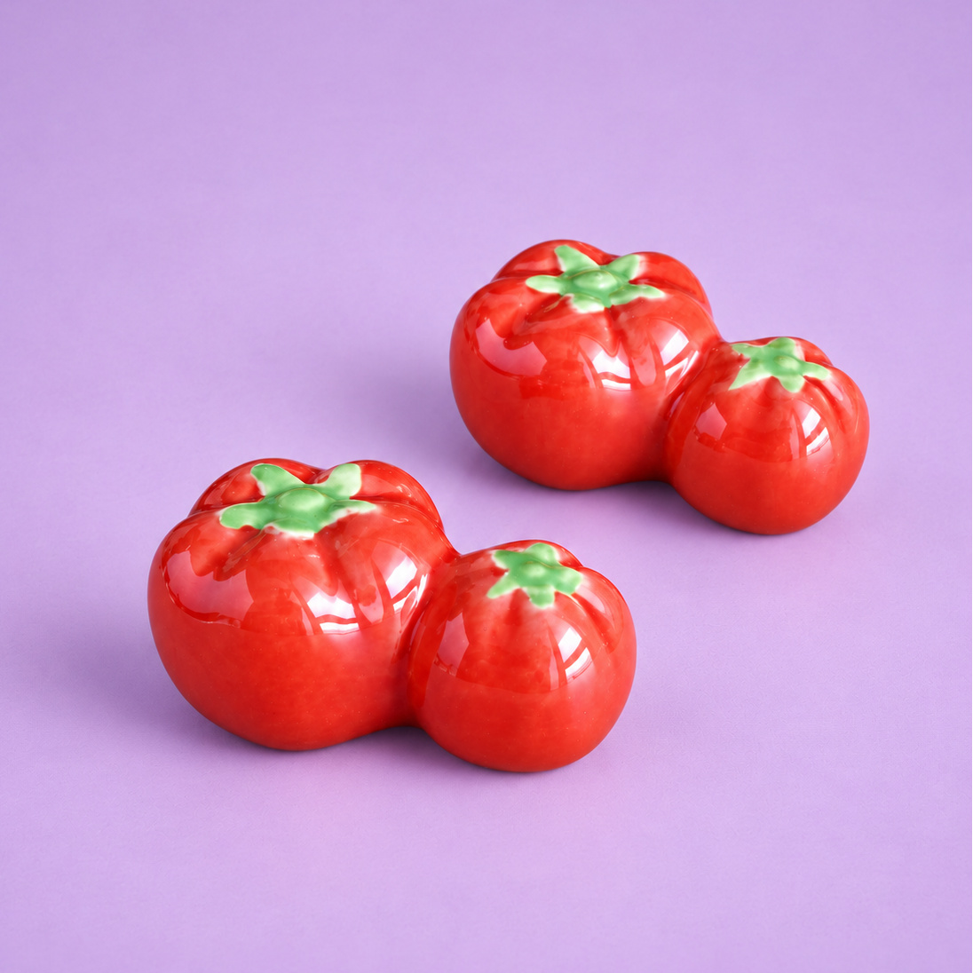 Three glossy red tomato-shaped ceramic decor or chopstick rest with green tops arranged on a solid lavender background.