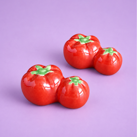 Three glossy red tomato-shaped ceramic decor or chopstick rest with green tops arranged on a solid lavender background.