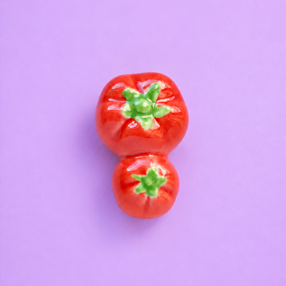 Two glossy red tomato-shaped ceramic ceramic decor or chopstick rest stacked vertically, set against a solid light purple background.