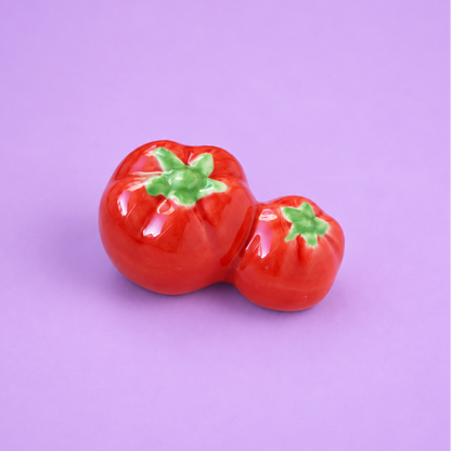 Two glossy red tomato-shaped ceramic ceramic decor or chopstick rest with green stem details, set against a solid light purple background.