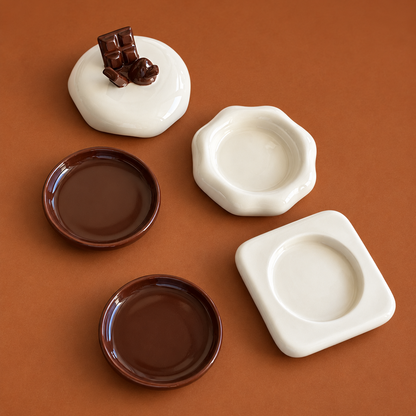Four small ceramic dish holders or coasters in white and dark brown, arranged on a warm brown background. One white piece is styled with a small chocolate chunk on top.