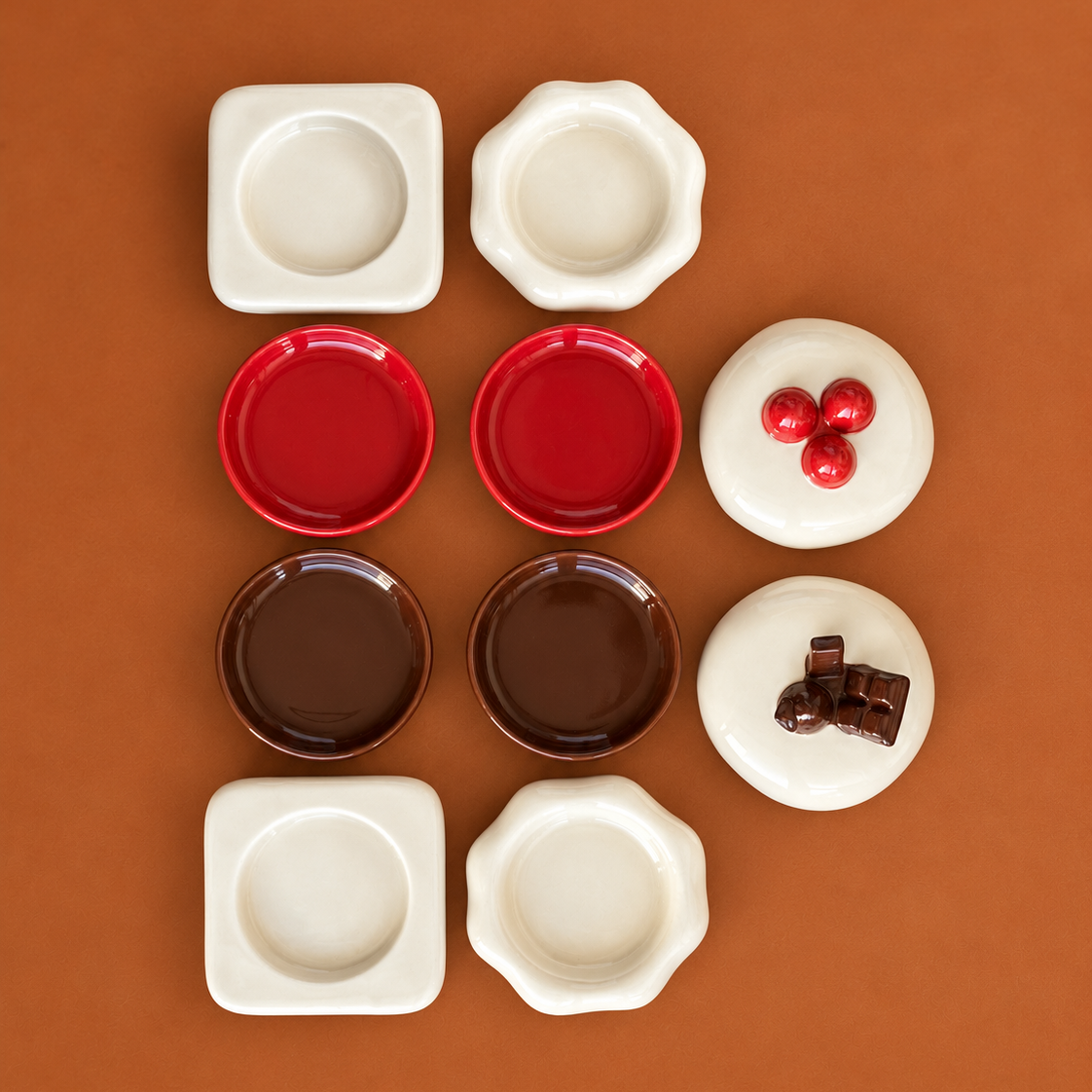 An overhead flat-lay of ceramic dipping bowls and small plates in multiple colors, including white, red, brown, and cream, arranged on a solid warm brown background. Some white pieces are decorated with red berries and dark brown pieces with chocolate chunks.