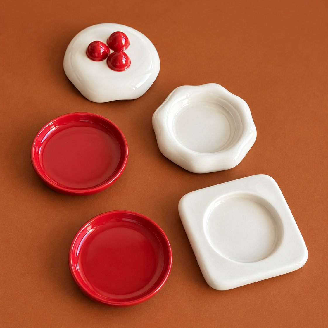 Four ceramic coasters in white and red arranged on a warm brown background. One white coaster has three raised red cherries, and two red coasters are shallow round dishes while one white coaster has a square border.