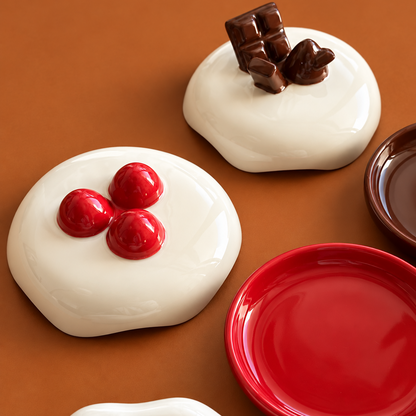 A set of glossy ceramic plates shaped like dessert toppings, including white cream with red cherries and chocolate chunks, a plain red plate, and a white plate with a red rim, arranged on a warm brown background.