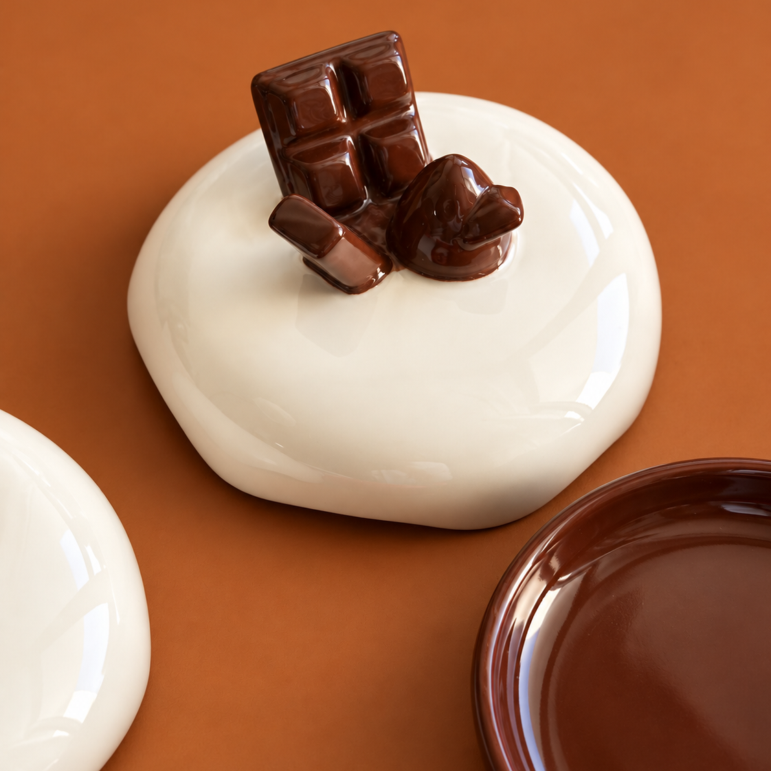 A glossy ceramic dish shaped like a white chocolate cookie topped with a dark chocolate bar and heart, set against a warm brown background with partial views of other colored dishes.