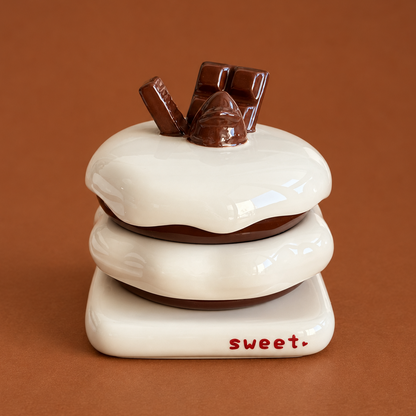 A ceramic s'mores jar or dish set designed to look like a stacked s'mores sandwich with white chocolate, brown chocolate, and dark chocolate pieces on top. The base is white ceramic with the word 'sweet.