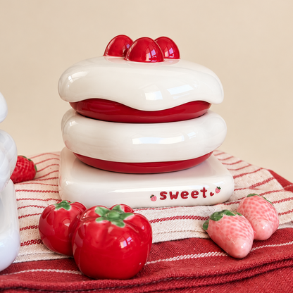 A ceramic dessert-themed container or figurine shaped like a red velvet cake with white frosting and red cherries on top, displayed on a red-and-white striped cloth with small strawberry and raspberry props against a warm beige background.