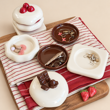 A styled flat-lay of ceramic jewelry organizers and trays with strawberry and chocolate motifs, arranged on a striped red and white cloth over a wooden board with small jewelry pieces placed inside.