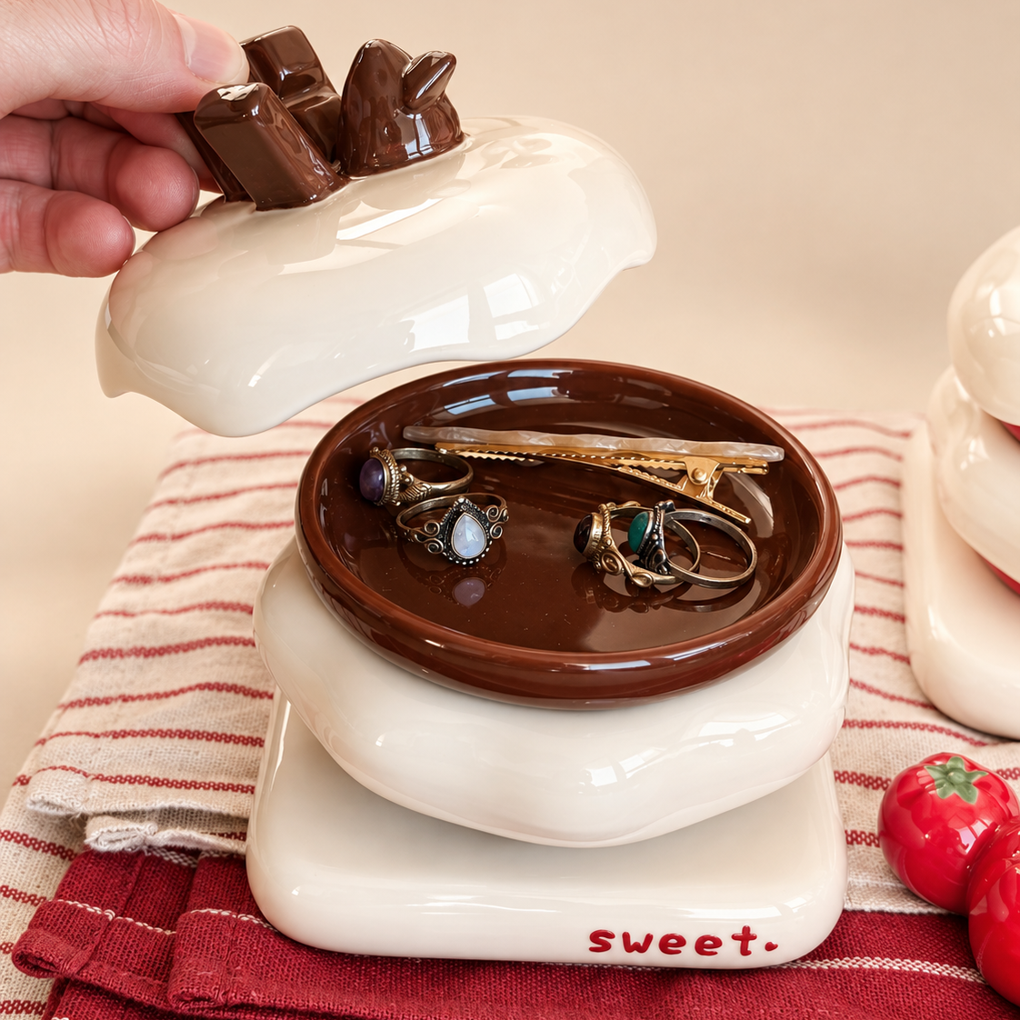 A ceramic jewelry box with a lid shaped like a chocolate bar, shown with a hand lifting the lid to reveal a brown interior tray holding earrings and a hair clip. The box sits on a red-and-white striped cloth with small strawberry decorations nearby.