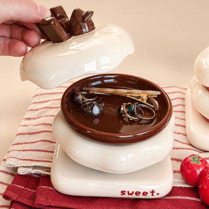 A ceramic jewelry box with a lid shaped like a chocolate bar, shown with a hand lifting the lid to reveal a brown interior tray holding earrings and a hair clip. The box sits on a red-and-white striped cloth with small strawberry decorations nearby.