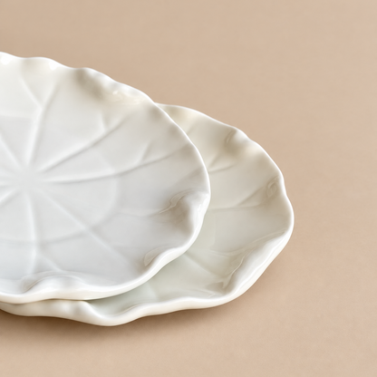 Two overlapping white ceramic plates with scalloped edges and a leaf-like embossed pattern, set against a solid beige background.